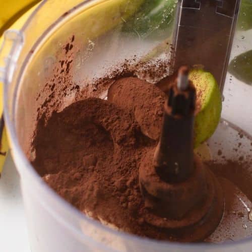 Adding cocoa powder to the Avocado Banana Pudding ingredients in a food processor.